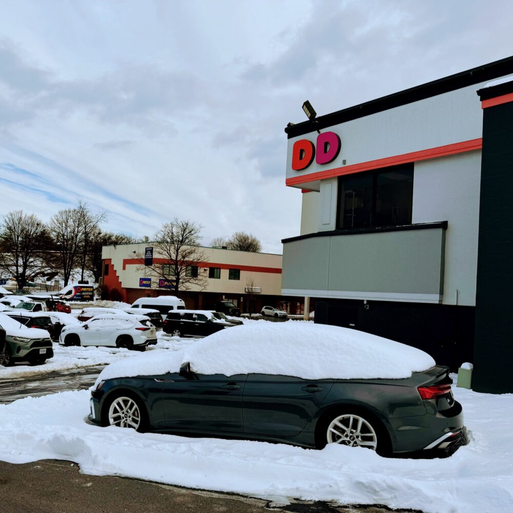 A snow covered car and parking lot at the donut shop.