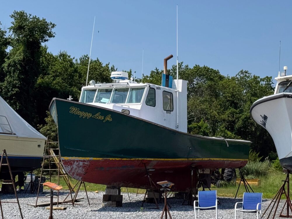 Green boat with a white cabin on blocks at a boatyard.